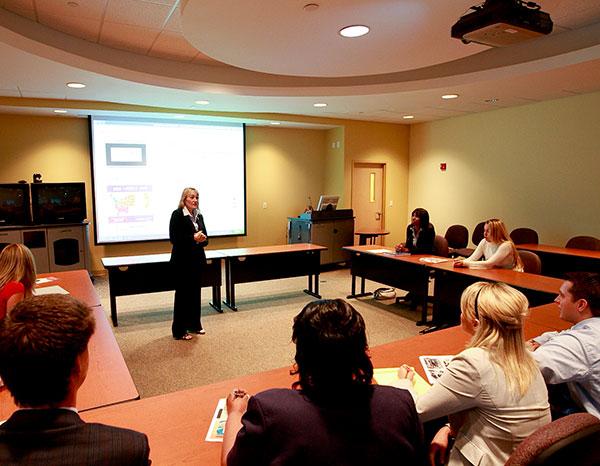 An adult woman speaks to a room full of adult students at tables arranged in a U shape around the room.