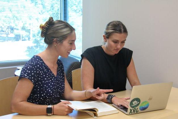 Two female graduate students talking and gesturing to one another while both looking a laptop screen..