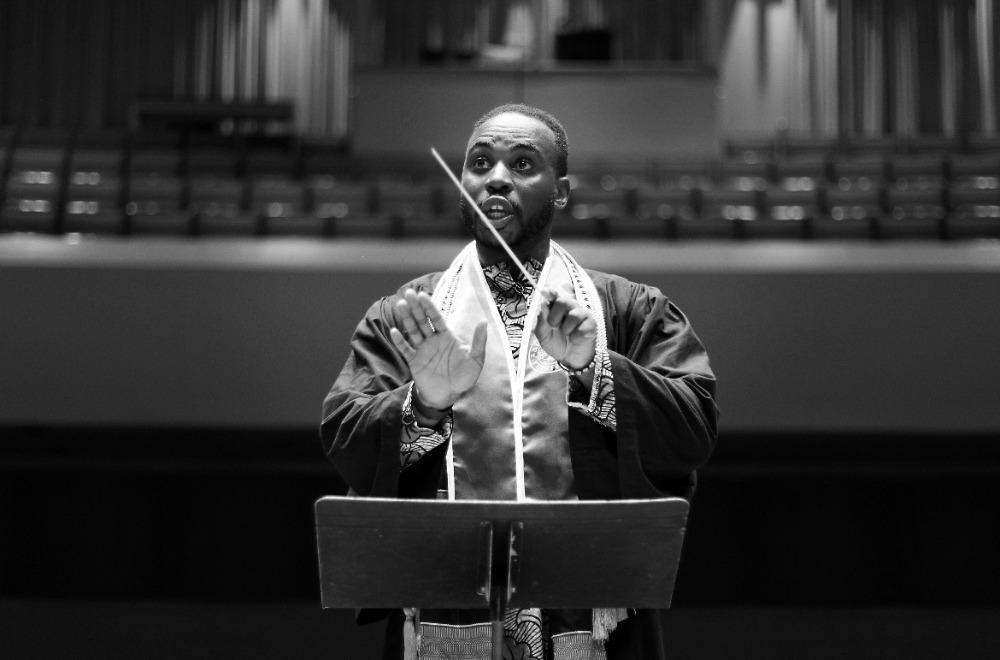 A black and white photo of a Black male student conductor wearing graduation regalia conducting an ensemble.