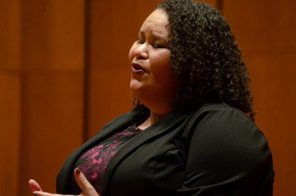 female student with dark curly hair in a dark suit and dress singing emotively 