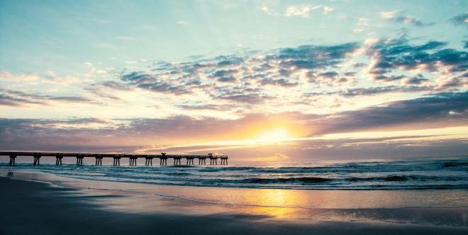 a picture of a beautiful sunrise over the beach next to the Jacksonville pier. The sky has blues, purples, and golds.