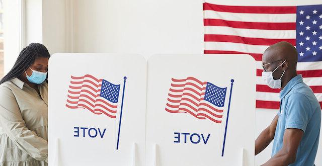 Photo of two people at voting stations with masks on placing their vote for an election with American flags on the voting booth and behind them