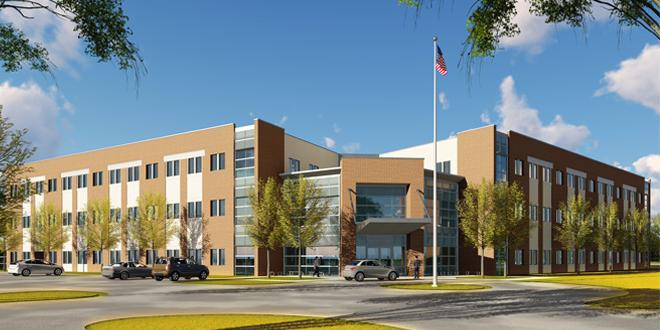 picture of a new building with tan and cream brick, three floors, and a number of windows. A cement roundabout is pictured at the bottom of the photo, and the American flag is at the top of the photo against a bright blue sky and a few clouds. 