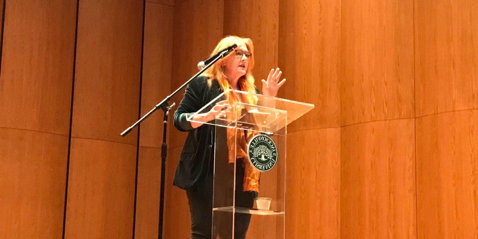 Caucasian female with blonde hair dressed in all black speaking emotively in front of a JU podium