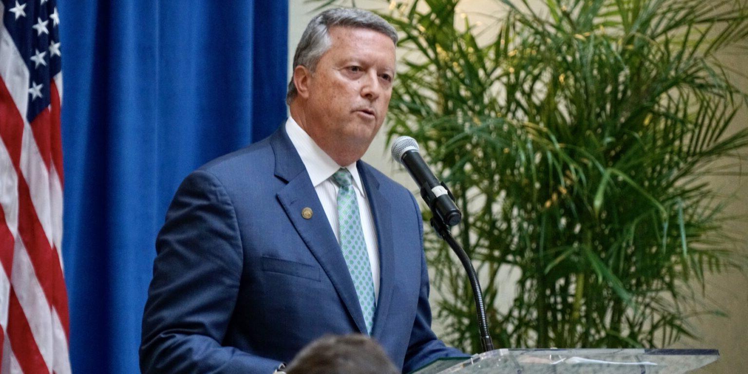 President of Jacksonville University with a green tie and blue suit in front of a podium speaking. There is an American flag to the right, and to the left is an artificial palm. 